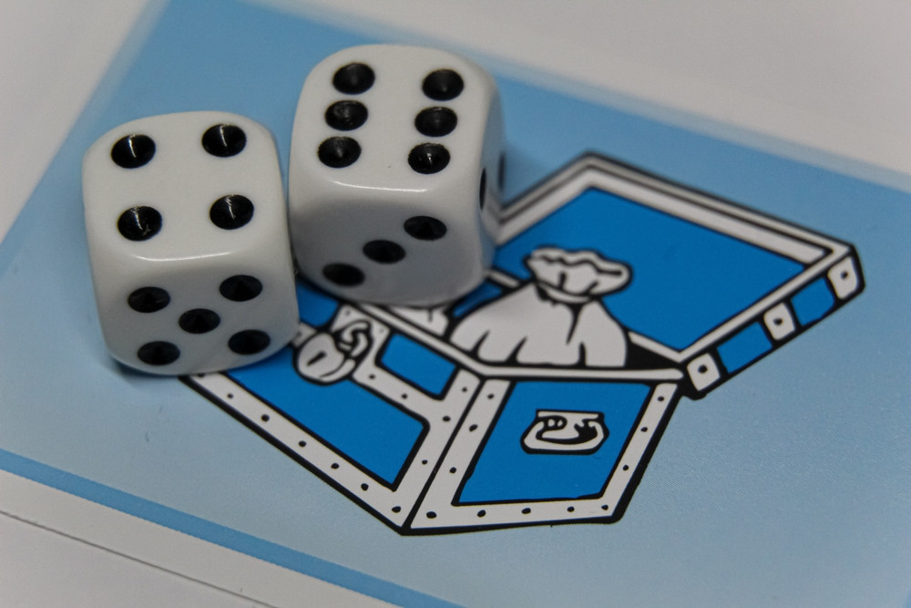 Two white dice with black dots are resting on a blue game card featuring a treasure chest with a bag inside.