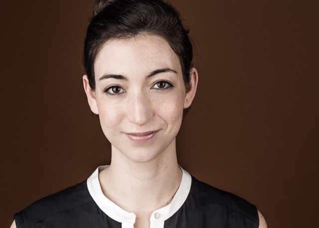 A woman with dark hair pulled back smiles softly against a warm brown background. She wears a black sleeveless top with a white collar.