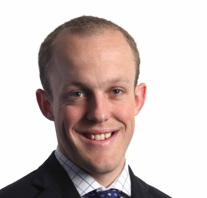 A smiling man wearing a dark suit and a checkered shirt, with short light brown hair and a confident expression.