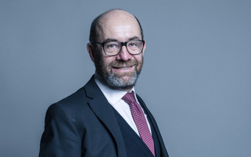 A bespectacled man with a beard smiles while wearing a suit and a tie, posing against a light gray background.