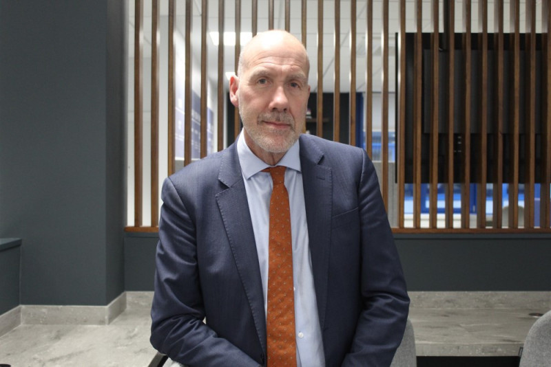 A man in a suit with a blue shirt and orange tie stands in an office setting, with wooden slat walls in the background. He has a serious expression.