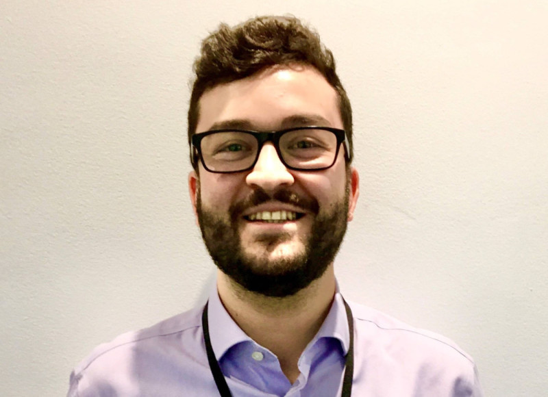 A smiling man with glasses and curly hair, wearing a light purple shirt and a dark necktie, stands against a light-colored wall.