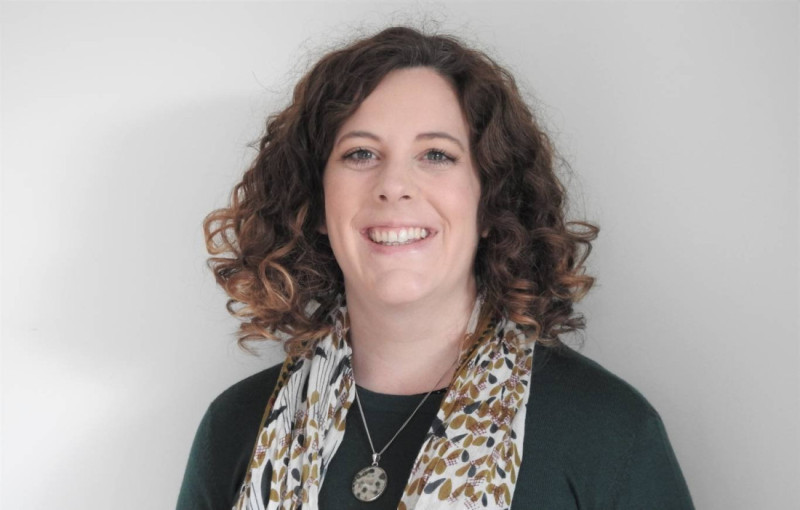 A woman with curly hair smiles warmly, wearing a green top and a patterned scarf around her neck. The background is light and neutral.