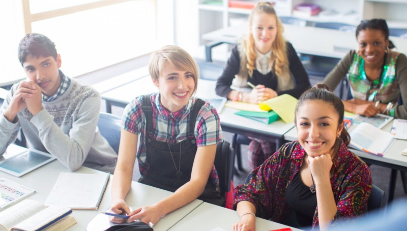 A diverse group of smiling students sitting at desks in a classroom, engaging with their studies. They have various books and notebooks in front of them, with some looking directly at the camera and others interacting with each other.