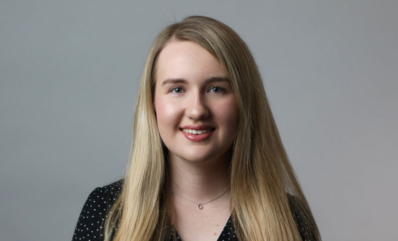 A smiling young woman with long, straight blonde hair wearing a black top with white polka dots, against a neutral gray background.