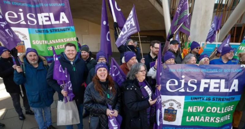 A group of people stands outside a building, holding banners and flags in purple and white. They are smiling and appear to be participating in a rally or demonstration, promoting unity and strength for educational institutions in Scotland.