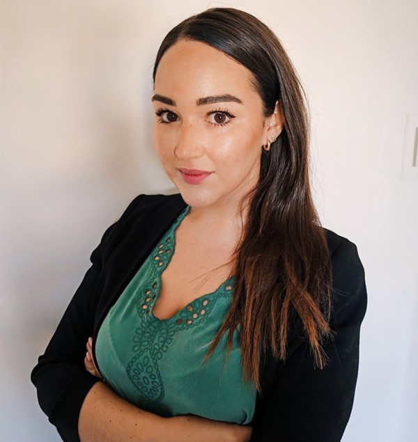 A woman with long, dark hair stands with her arms crossed, wearing a green top with decorative embroidery and a black blazer. She looks confidently at the camera against a plain background.