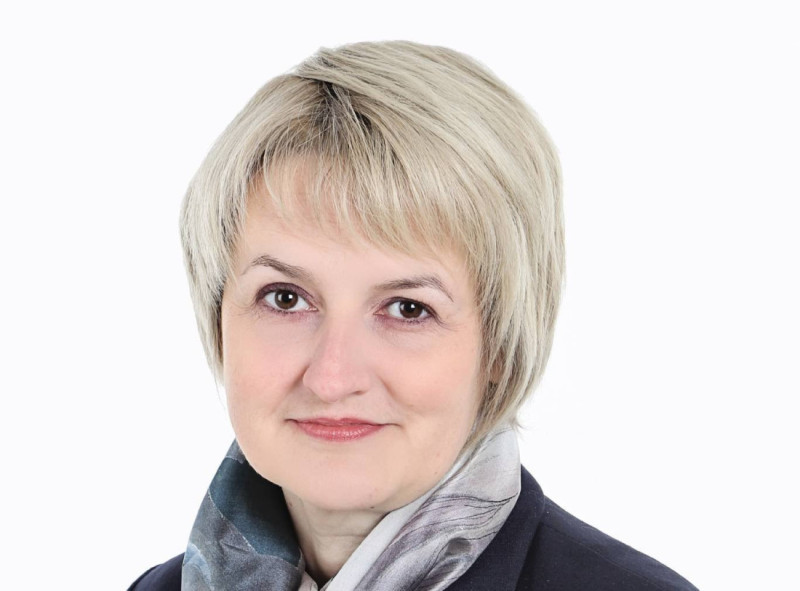 A woman with short, light blonde hair and a subtle smile, wearing a dark blazer and a patterned scarf. The background is plain white.