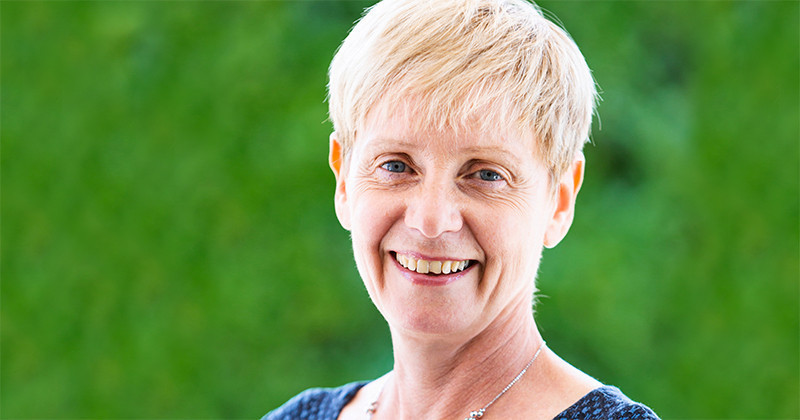 Smiling woman with short blonde hair, wearing a navy top, set against a blurred green background.