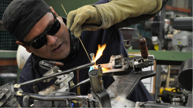 A technician wearing safety gloves and glasses is welding metal components with a flame, focused on his work in a workshop environment.