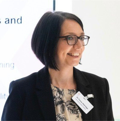 A woman with short dark hair and glasses smiles while speaking at a presentation. She is wearing a black blazer over a floral top and has a name badge clipped to her blazer.