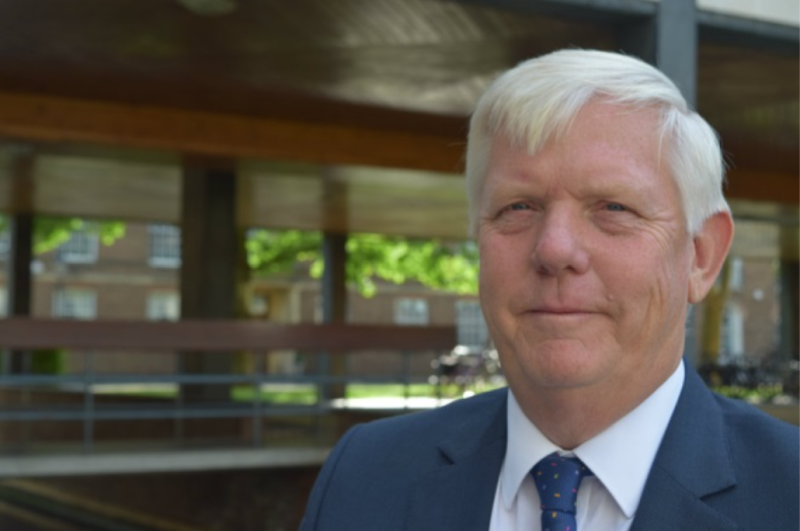 A man with short white hair and a blue suit stands outdoors, smiling slightly. He is in front of a modern wooden structure with greenery in the background.