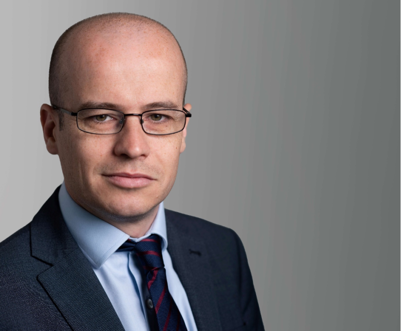 A confident man in a dark suit and tie stands against a gray background, wearing glasses and looking directly at the camera. His bald head and serious expression convey professionalism.