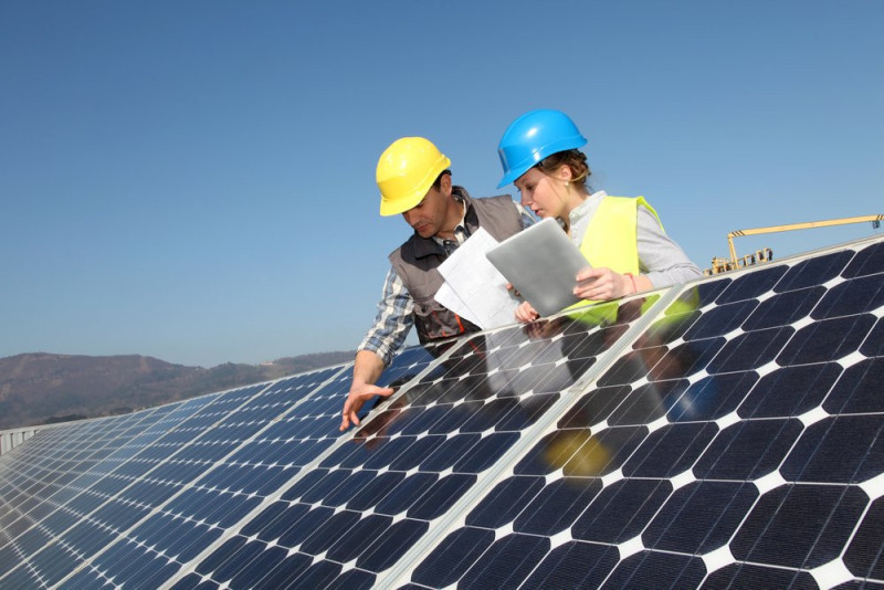 Two workers in hard hats and safety vests inspect solar panels on a rooftop. One is holding a tablet while the other points at the panels, with a clear blue sky in the background.