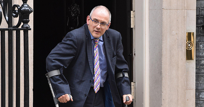 A man in a suit with glasses is exiting a building using crutches. He has a surprised expression and is wearing a colorful tie.