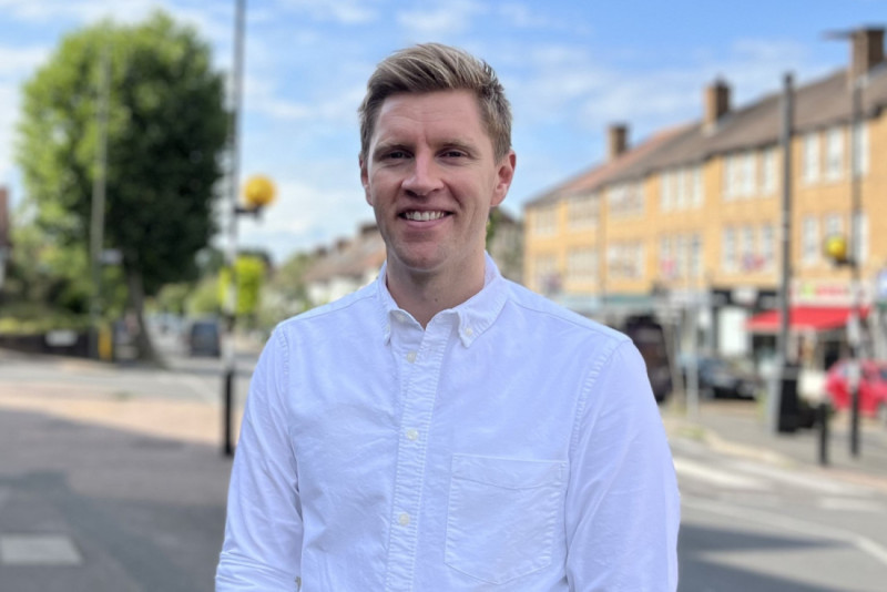 A young man with short blonde hair smiles while standing on a street lined with brick buildings and trees under a clear blue sky. He is wearing a white button-up shirt.