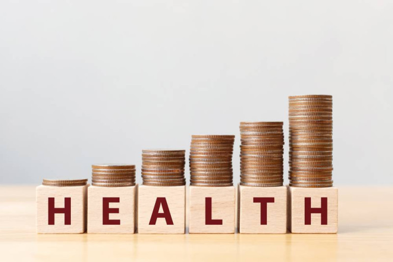 Stacked coins of increasing height next to wooden blocks spelling "HEALTH." The arrangement symbolizes the connection between health and wealth.