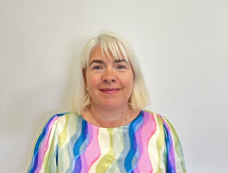 A woman with shoulder-length blonde hair and wearing a colorful striped blouse smiles at the camera against a plain white background.