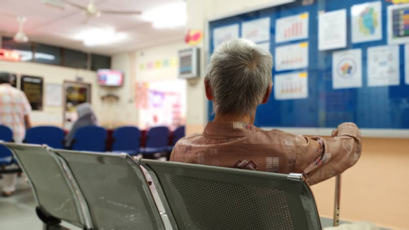 An elderly man sits in a waiting area, facing away from the camera. He wears a patterned shirt and holds a cane, with various posters and charts displayed on the wall behind him.