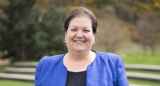 A woman with dark hair and pearls smiles warmly while standing outdoors in a park, wearing a blue blazer over a black top. Trees and grassy areas are visible in the background.