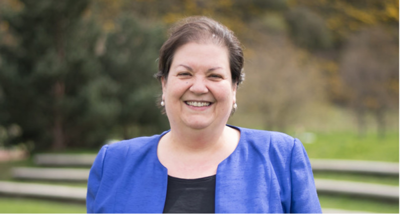 A smiling woman with dark hair wearing a blue blazer stands outdoors, surrounded by greenery.