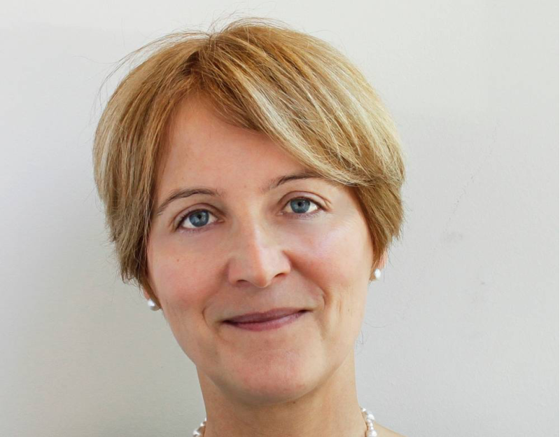 A woman with short blonde hair smiles at the camera, wearing a pearl necklace and a subtle expression. She has blue eyes and a light complexion, with a neutral background.