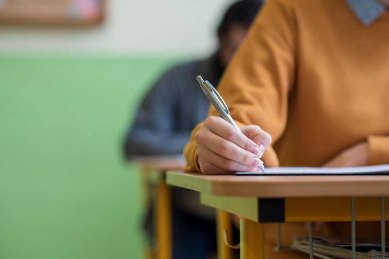 A student in a brown sweater writes in a notebook at a desk, with a blurred classmate visible in the background.