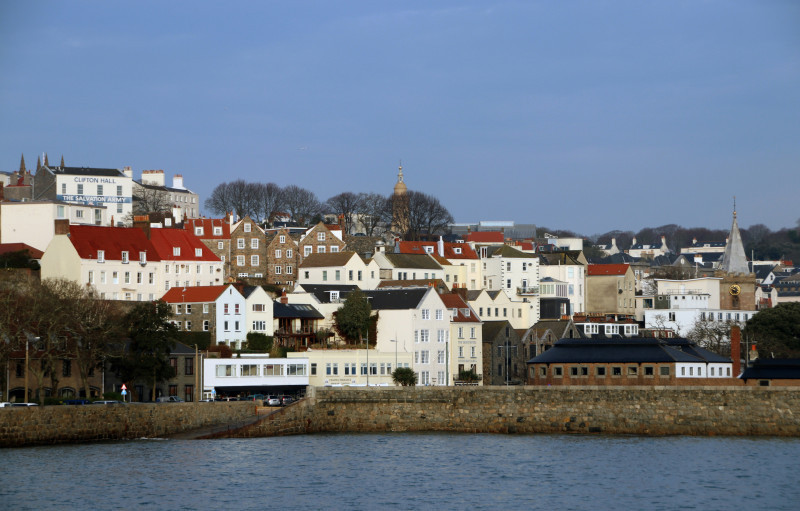 Coastal view of a picturesque town with a mix of historic and modern buildings, featuring red-tiled roofs and a clear blue sky in the background.