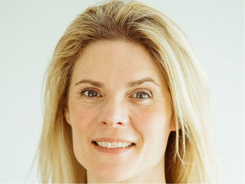 A woman with long, light blonde hair smiles gently, standing in front of a light-colored background. Her expression is warm and approachable, with subtle makeup accentuating her features.