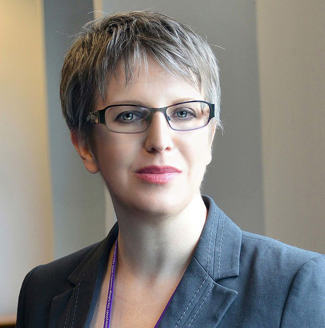 A woman with short, silver hair and glasses smiles confidently while wearing a blazer. She has a professional demeanor and is standing indoors.