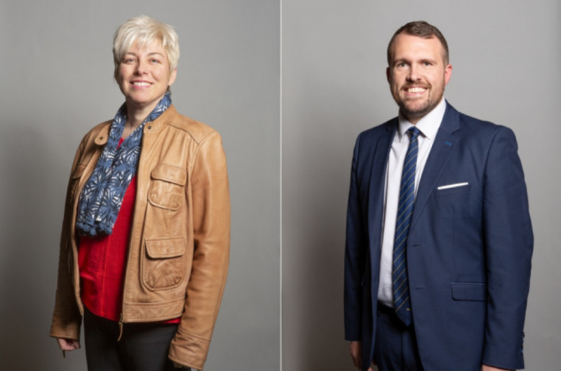 Two individuals are standing against a neutral grey background. On the left, a woman with short blonde hair wears a brown leather jacket and a blue scarf over a red shirt. On the right, a man with short brown hair is dressed in a navy suit with a light blue shirt and a patterned tie, smiling confidently.