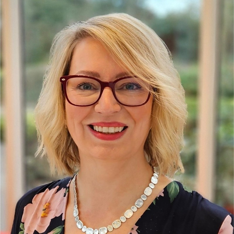 A smiling woman with blonde, shoulder-length hair and red glasses wears a floral top and a silver necklace. She is indoors with greenery visible in the background.