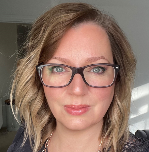 A woman with shoulder-length wavy hair and glasses is looking directly at the camera, smiling softly. She is wearing a black top and has a natural makeup look, with light pink lipstick. Sunlight casts a warm glow on her face.