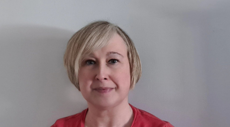 A woman with short blonde hair wearing a red shirt stands against a plain white wall, looking directly at the camera.