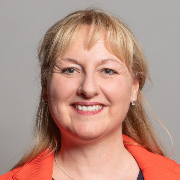 A smiling woman with blonde hair and light skin is wearing an orange blazer. She has a friendly expression and is facing the camera.