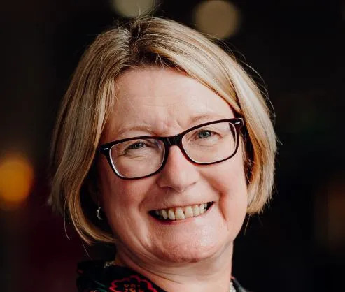 A smiling woman with short blonde hair and glasses, dressed in a colorful patterned top, stands against a blurred background.