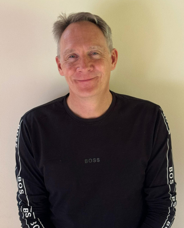 A smiling man stands against a plain wall, wearing a black long-sleeve shirt with "BOSS" printed on it.