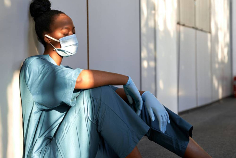 A healthcare worker wearing a blue medical gown, a surgical mask, and gloves sits against a wall, appearing contemplative with closed eyes, in a softly lit environment.