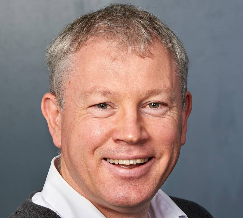 A smiling man with short gray hair and a light complexion, wearing a white shirt and a dark sweater, poses against a gray background.