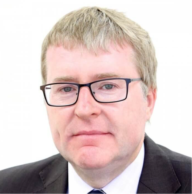 A man with short, light brown hair and glasses is wearing a dark suit and tie. He has a neutral expression and is set against a plain white background.