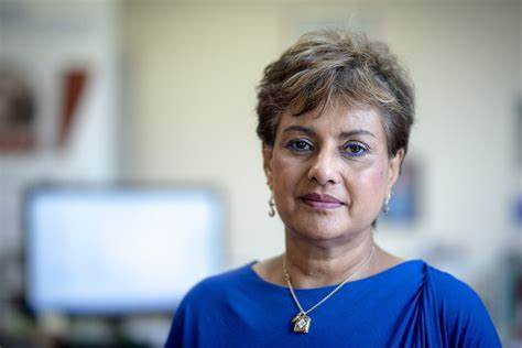 A woman with short, styled hair and light makeup is wearing a blue top. She has a neutral expression and is standing in a softly lit environment.