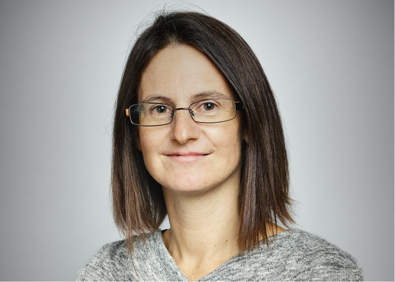 A woman with shoulder-length brown hair, wearing glasses and a gray sweater, smiles gently against a light gray background.