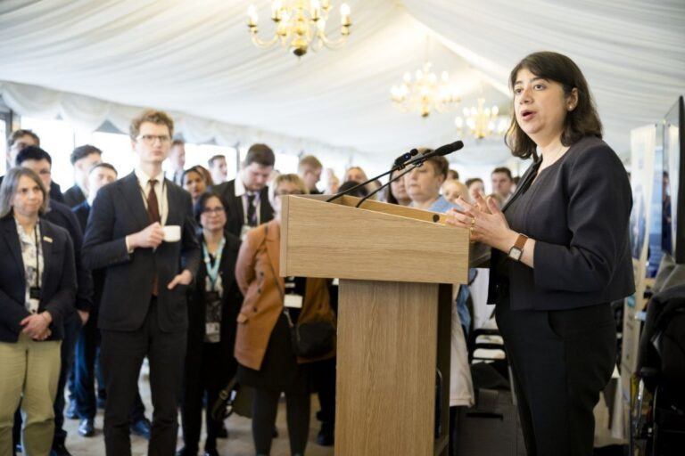 A woman speaks at a podium in front of a large audience in a tented setup. She gestures while engaging with the crowd, which appears to be listening intently.