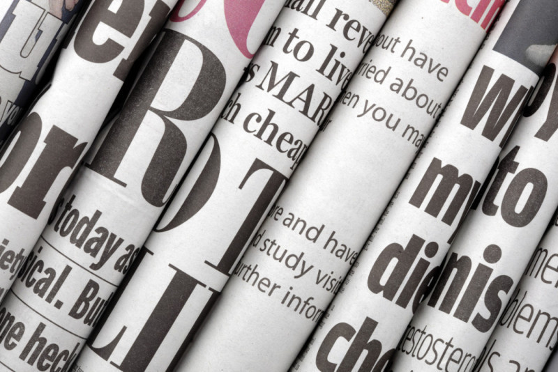 Close-up of rolled newspapers with various headlines and text. The papers feature large, bold typography in black and red, suggesting a variety of topics and news.