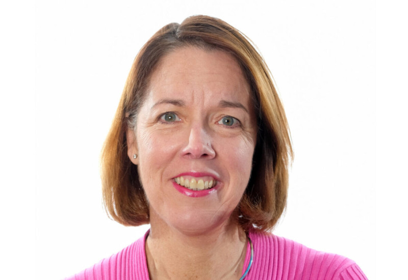 A woman with shoulder-length brown hair smiles at the camera, wearing a bright pink sweater. She has light makeup and is set against a plain white background.