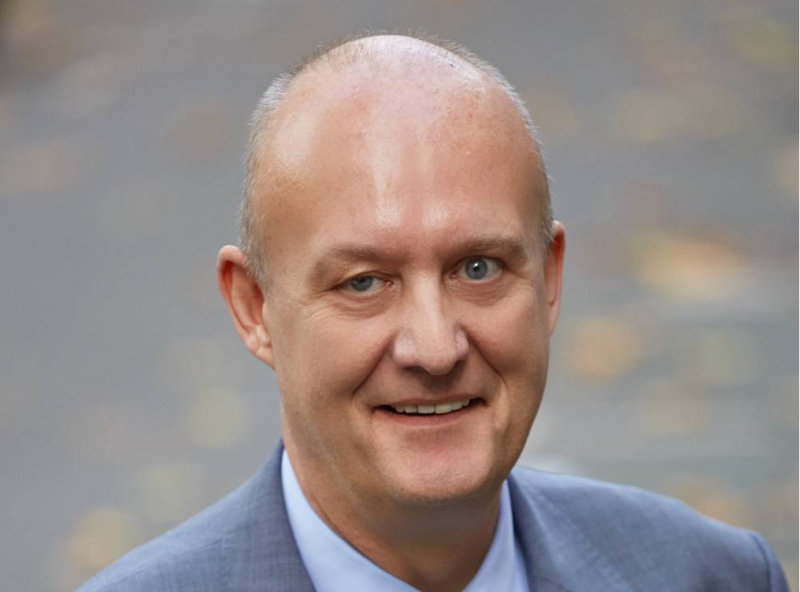 A bald man with a friendly smile wears a gray suit and a light blue shirt. He is outdoors, with a blurred background of autumn leaves on the ground.