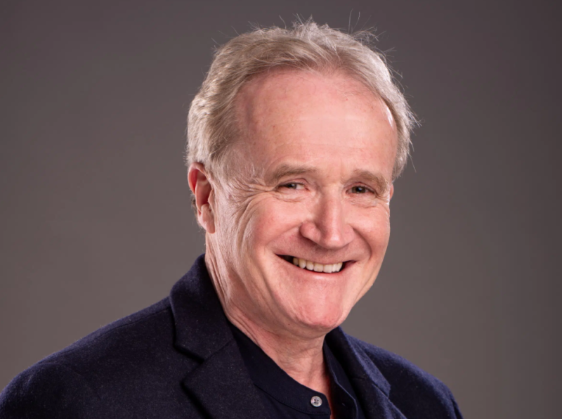 A smiling man with gray hair, wearing a dark blazer over a collared shirt, poses against a neutral background.