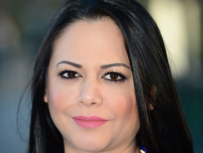 A woman with long black hair and warm brown eyes smiles gently, wearing subtle makeup and a light pink lip color.