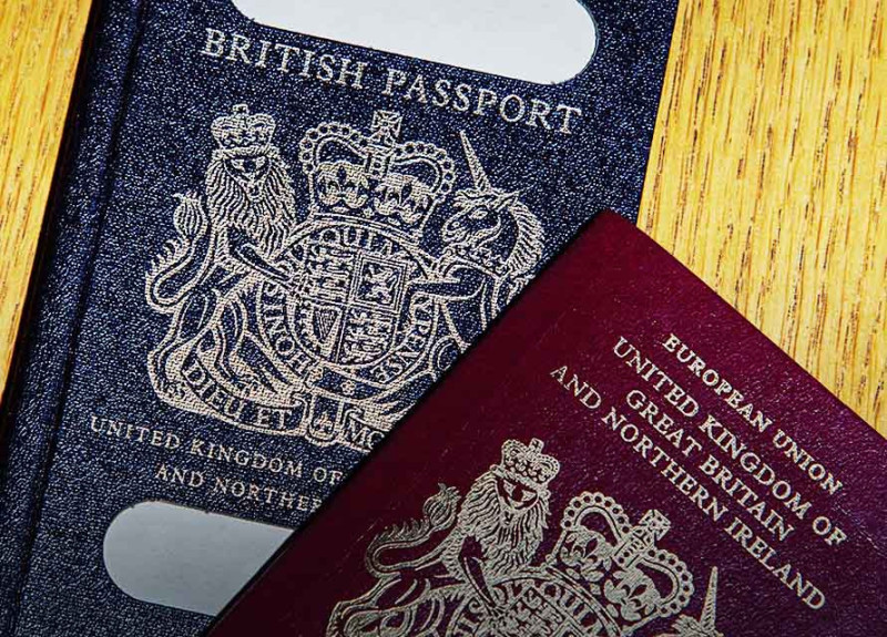 Two British passports are displayed on a wooden surface. One is dark blue, featuring the golden crest of the United Kingdom, while the other is maroon with a similar crest, labeled as European Union.