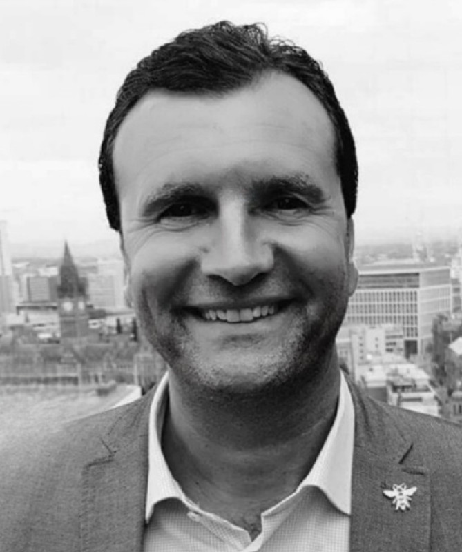 A man in a light gray suit smiles at the camera, with a city skyline visible in the background. He has dark hair and wears a pin on his lapel. The image is in black and white.